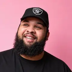 Tom Koko Bikusa, souriant, en casquette noire sur fond rose